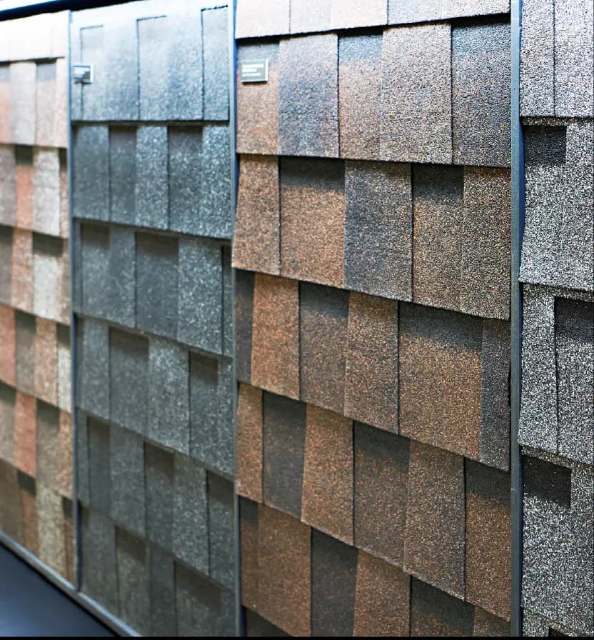 Display wall showing variety of roofing shingle samples in different colors including brown tan gray and black textures Mobile