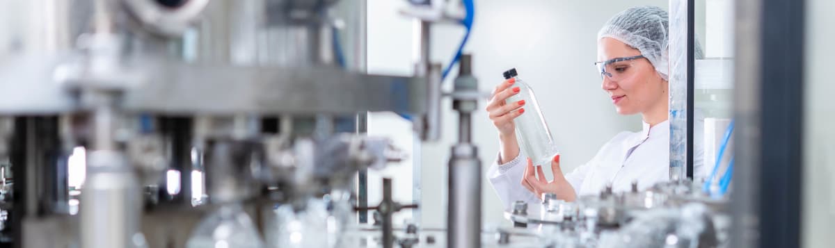 Female quality control technician in hairnet and safety glasses inspecting clear bottle on automated manufacturing line
