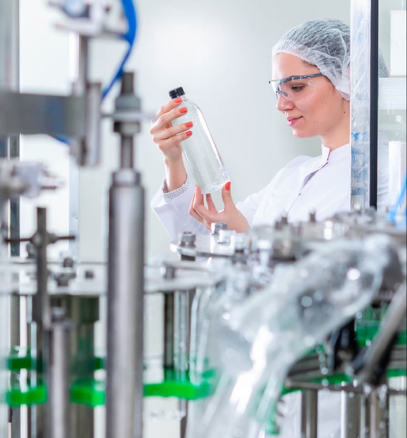 Female quality control technician in hairnet and safety glasses inspecting clear bottle on automated manufacturing line Mobile