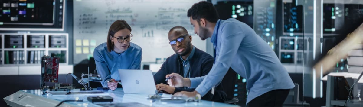 Diverse technology team collaborating on laptop with circuit boards in modern command center control room workspace