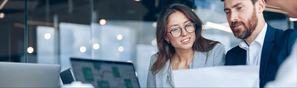 Smiling business woman wearing glasses and male colleague collaborating on laptop computer in contemporary office workspace environment